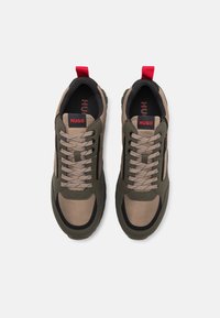 Las zapatillas tienen un upper de malla en tonos marrón y verde oliva con detalles en negro, cordones planos, lengua acolchada y una pestaña roja en el talón.