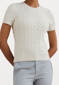 Kurzärmliger, cremefarbener Strickpullover mit Kabelmuster, gerippten Bündchen und Saum, der ein strukturiertes Muster und eine taillierte Passform aufweist. Über hellblauen Hosen getragen.