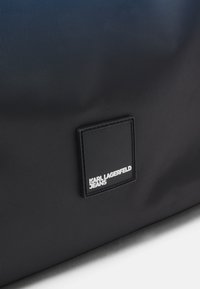 Saco de tecido preto com textura suave, design retangular e um destacado emblema preto com texto branco, "KARL LAGERFELD JEANS."