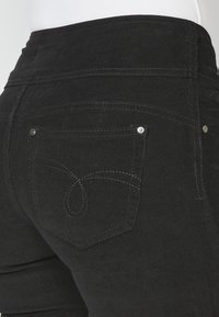 Pantalon en velours côtelé noir avec une poche arrière présentant des surpiqûres décoratives et des rivets métalliques, visible en gros plan de profil arrière.