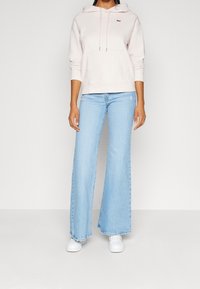 Haut à capuche rose clair avec poche avant et capuche cordée, associé à un jean bleu clair à jambes larges et des baskets blanches. Tenue simple et décontractée.