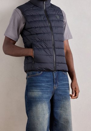 Homme portant une veste matelassée noire par-dessus une chemise grise à manches courtes, une main dans la poche de la veste et un jean bleu.
