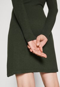Abito lavorato a maglia verde scuro con maniche lunghe, un corpino aderente e un orlo svasato. Le mani sono posizionate dietro la schiena, evidenziando la texture del tessuto.