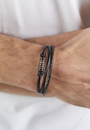 Man's wrist wearing three black leather bracelets, one with a cylindrical bead, against a white shirt background.