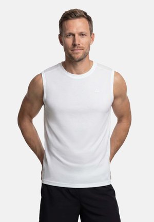 Homme aux cheveux courts portant un débardeur blanc de sport et un short noir, debout les mains dans le dos.