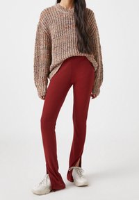 Maglione marrone lavorato a maglia con una vestibilità morbida, abbinato a pantaloni bordeaux aderenti con spacchi laterali e sneakers bianche.