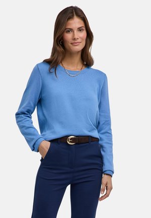 Frau mit langen braunen Haaren, die ein hellblaues Langarmshirt, dunkelblaue Hose, braunen Gürtel und eine goldene Halskette trägt und mit einer Hand in der Tasche steht.