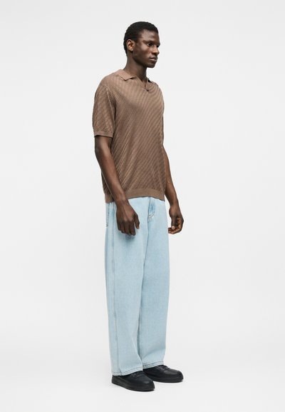 Homme debout, portant une chemise marron à manches courtes texturée, un jean large bleu clair et des chaussures noires, sur un fond blanc uni.