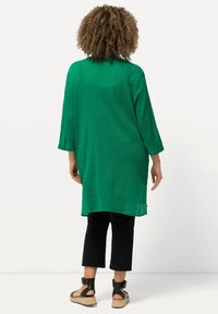 Túnica verde, holgada, con mangas de tres cuartos, hecha de un tejido texturizado. Combinada con pantalones negros cortos y sandalias negras con tiras.