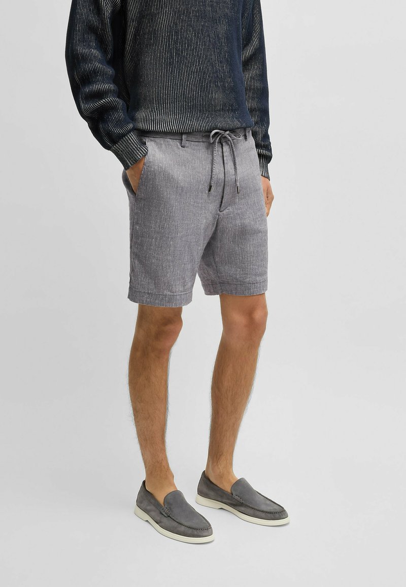 Vaaleanharmaat shortsit, jotka on valmistettu teksturoidusta kankaasta. Niissä on kiristysnauha tummilla yksityiskohdilla vyötäröllä. Yhdistetty harmaisiin liukuvasti jalkaan laitettaviin kenkiin.