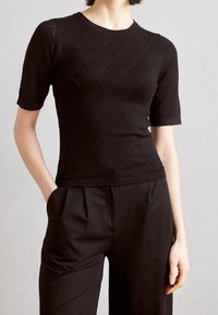 Kvinna iklädd en figursydd svart kortärmad stickad topp med diamantmönster och svarta högmidjade byxor med veck och fickor.