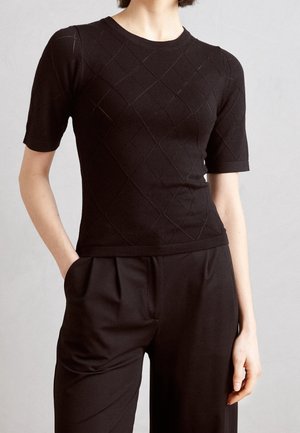 Femme portant un haut en tricot noir moulant à manches courtes avec un motif en losange et un pantalon noir taille haute avec des plis et des poches.