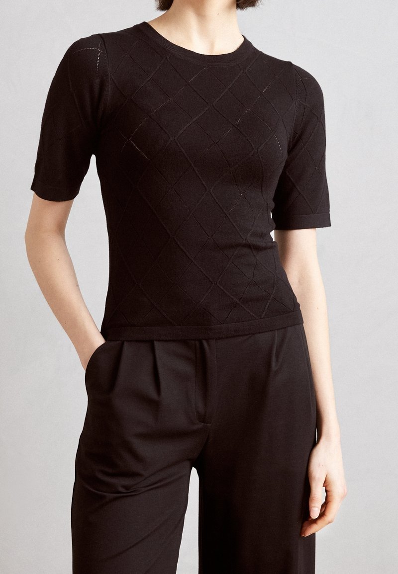 Kvinna iklädd en figursydd svart kortärmad stickad topp med diamantmönster och svarta högmidjade byxor med veck och fickor.