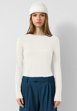 Jeune femme portant un haut blanc à manches longues, un pantalon plissé bleu foncé et un bonnet blanc en tricot, debout devant un fond clair uni.
