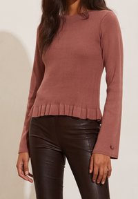 Maglione a coste rosa con maniche lunghe e orlo a volant, abbinato a pantaloni aderenti marroni lucidi. Il tessuto è liscio e aderente.