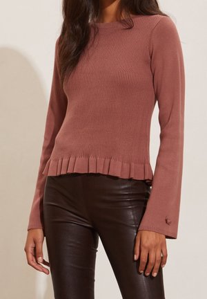 Rosa gerippter Pullover mit langen Ärmeln und gerafftem Saum, kombiniert mit glänzenden, braunen, figurbetonten Hosen. Der Stoff ist glatt und figurbetont.