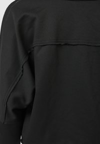 Svart plagg med ett mjukt, texturerat tyg. Har en rå kant längs sömmen och en lös passform. Designen betonar enkelhet och funktionalitet.