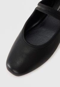 Musta nahkainen ballerinatossu pyöreällä varvastyllä, jossa on teksturoitu pohjallinen ja kapea elastinen hihna jalkapöydän ympäri. Tasainen pinta.