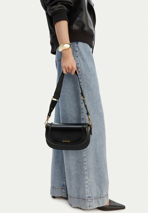 Persona con vaqueros de pierna ancha de color azul claro, chaqueta de cuero negra, pulsera dorada y sosteniendo un pequeño bolso negro Badura con correa ajustable.