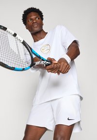 Hvit t-skjorte med kort erme fra Nike med et gyllent logo, parret med matchende hvite shorts. Spilleren holder et blått og svart tennisracket.