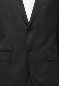 Κοντινή λήψη σκούρου καφέ σακακιού με δύο μαύρα κουμπιά κουμπωμένα πάνω από λευκό πουκάμισο, με δύο τσέπες με φλά π στην πρόσοψη.