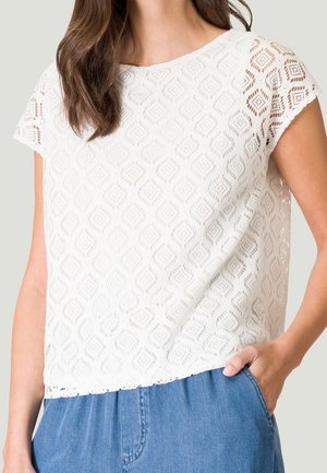 Top in pizzo bianco con un motivo geometrico, maniche corte e orlo a scalloppatura, abbinato a pantaloni in denim azzurro chiaro.