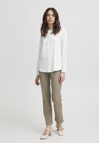 Camicia bianca a maniche lunghe con colletto, frontale con bottoni, abbinata a pantaloni beige slim-fit e scarpe con tacco blocco chiare. Tessuto liscio e linee pulite.