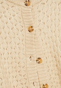 Cardigan en maille beige clair avec un motif en losange, un col côtelé et cinq boutons ronds en écaille de tortue sur le devant.
