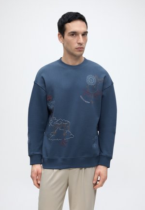 Mann trägt einen blauen Sweatshirt mit abstrakten bestickten Designs und beigen Hosen, steht vor einem einfarbigen hellen Hintergrund.