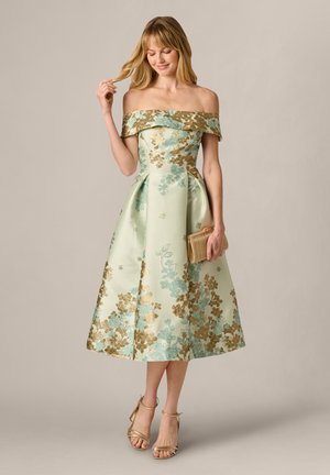 Žena v mintově zelených šatech midi délky s odkrytými rameny, zdobených zlatými a modrými květinovými vzory, držící zlaté psaníčko a nosící zlaté lodičky.