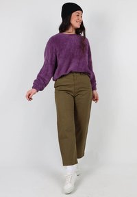 Un pull violet duveteux, un pantalon large olive, des chaussettes blanches et des baskets blanches, le tout complété par un bonnet. Le pull a une coupe ample et une surface texturée.