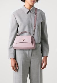 Jasnoróżowa skórzana torebka o strukturalnym designie, z górnym uchwytem i odpinanym paskiem. Wyposażona w złote okucia i trójkątne logo.
