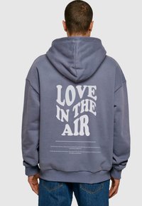 Grå hoodie med en stor hvid grafikteks "KÆRLIGHED I LUFTEN" på ryggen og yderligere tekst nedenfor. Blødt stof, afslappet pasform, hætte-design.