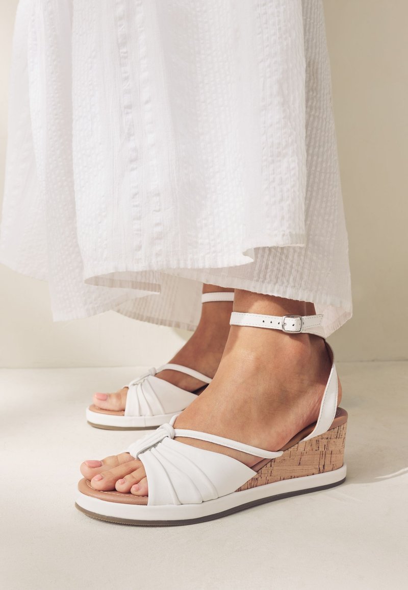 Sandales compensées blanches avec talon en liège, design plissé à l'avant et bride ajustable à la cheville. Jupe en tissu blanc texturé au-dessus.
