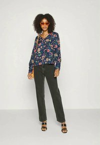 Blusa floral em azul marinho escuro com padrões vibrantes em rosa e verde, combinada com jeans escuros e sandálias pretas. A modelo seguro uma bolsa laranja.