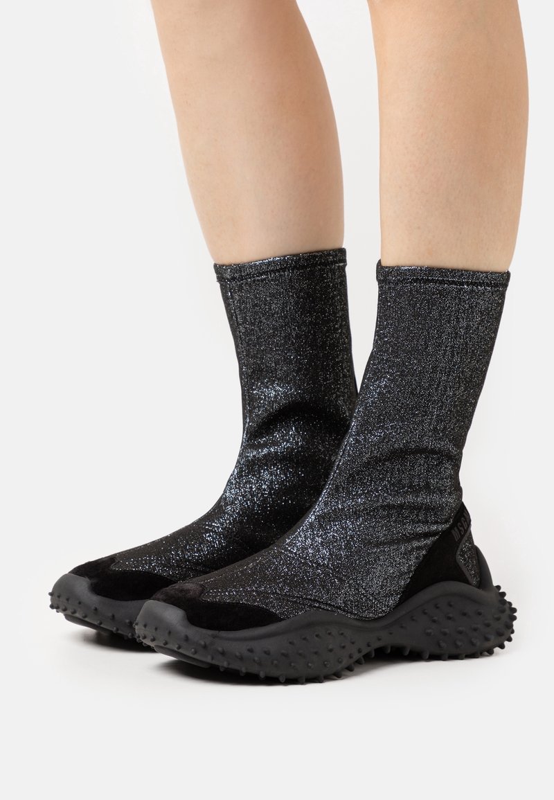 Bottes noires pailletées style chaussette avec semelles épaisses texturées portées sur des jambes, sur un fond clair uni.