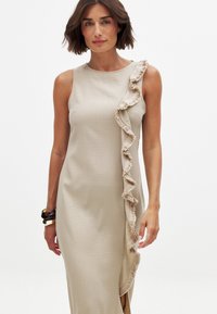 Beżowa, bezrękawnikowa sukienka z bocznym falbanką. Gładka tekstura, średniej długości krój, minimalistyczny styl oraz subtelne detale wzdłuż krawędzi falbanki.