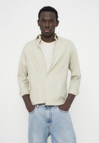 Ανοιχτό πράσινο πουκάμισο με απαλό ύφασμα, μανίκια γυρισμένα και ευθεία εφαρμογή, φορεμένο πάνω από ανοιχτό μπεζ πουλόβερ και μπλε τζιν.