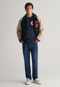 Námořnická varsity bunda s béžovými rukávy, červenými akcenty a logem; spárovaná s námořnickým tričkem, denimovými džíny a světlými teniskami.