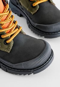 Bottes robustes noires et vert olive avec des embouts d'orteils épais côtelés et des lacets tressés jaune-orange sur une surface claire.