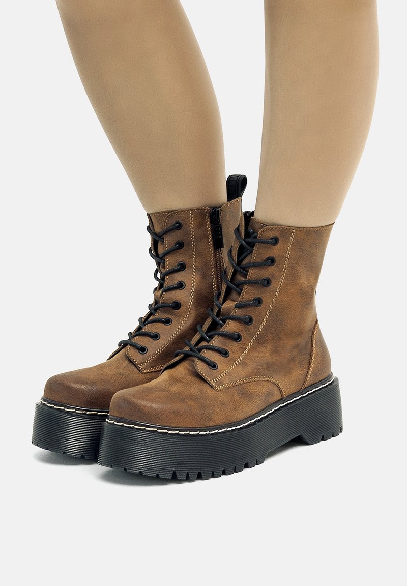 Bottines montantes en cuir marron avec semelles plateformes noires épaisses portées sur des jambes nues sur un fond clair.