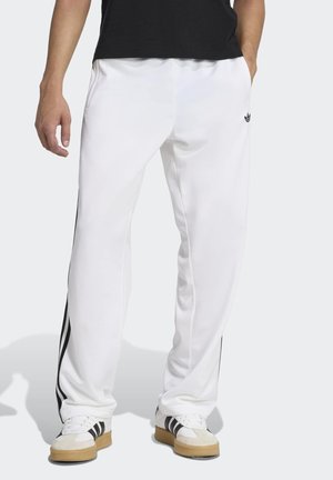 Mann trägt weiße Adidas-Trainingshose mit schwarzen Streifen an den Seiten und weiße Sneaker mit schwarzen Streifen, steht mit Händen in den Taschen.