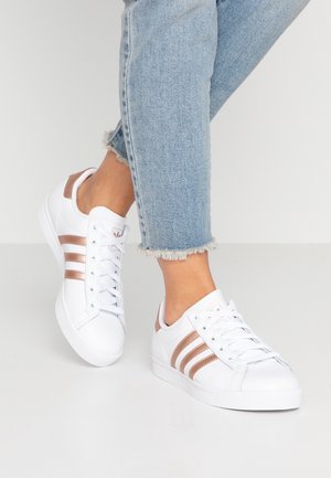 Osoba nosząca jasno niebieskie jeansy z postrzępionymi krawędziami i białe trampki z różowo-złotymi paskami, stojąca na białej podłodze.