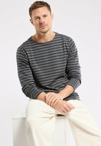 Maglietta a maniche lunghe, colletto rotondo, di colore verde oliva con righe orizzontali blu navy; indossata con pantaloni chiari e strutturati. Design semplice e casual.