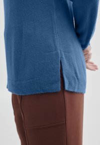 Blå stickad tröja med ribbad nederkant och sidoflikar, kombinerad med snäva bruna byxor. Texturerat material, avslappnad passform.