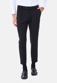 Pantaloni neri su misura con pieghe, chiusura con bottone e tasche laterali. Indossati con mocassini neri lucidi e calzini neri.