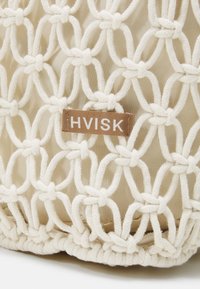 Sac fourre-tout en filet beige avec un design en corde tissée, comportant une étiquette marron avec "HVISK" en lettres blanches cousue sur le côté.