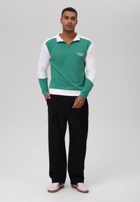 Pulóver de manga larga verde y blanco con cuello de cremallera, con un acabado texturizado y un logo en el pecho, combinado con pantalones negros.