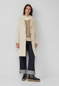 Frau in einem cremefarbenen Mantel, braunem und cremefarbenem Strickpullover, dunklen, weit geschnittenen Jeans und schwarzen, klobigen Stiefeln, die an einer grauen Wand steht.