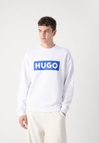 Baltas džemperis su mėlynu stačiakampiu logotipu, kuriame rašoma "HUGO" didžiosiomis raidėmis. Turi apvalų kaklą, ilgomis rankovėmis ir guma apdirbtus rankogalius.
